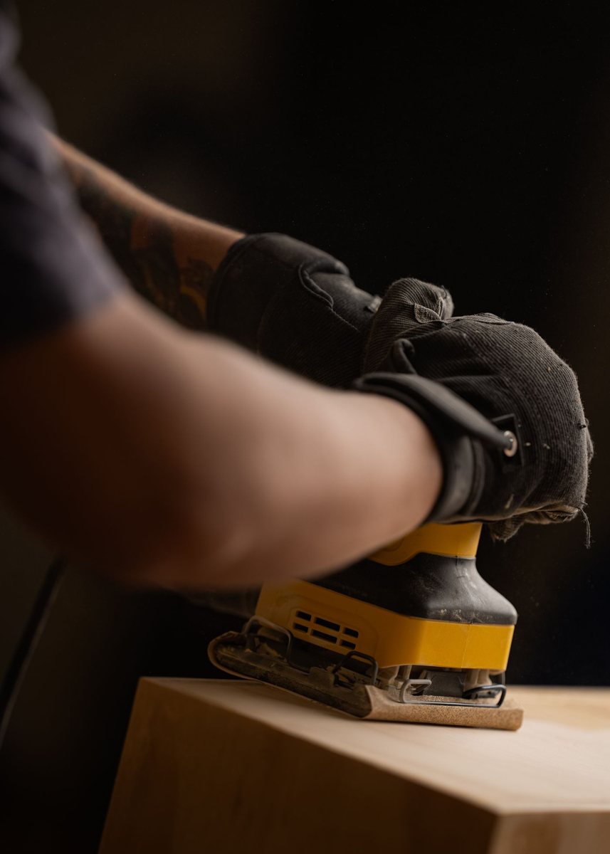 Gloved hand guiding a quarter-sheet palm sander along the edge of a board
